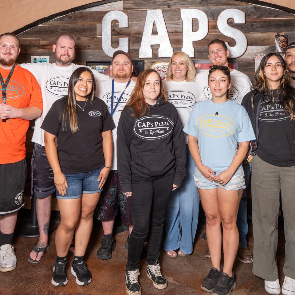 Smiling Cap's Pizza & Tap House staff in branded shirts stand before a wooden wall with 'CAPS' sign.