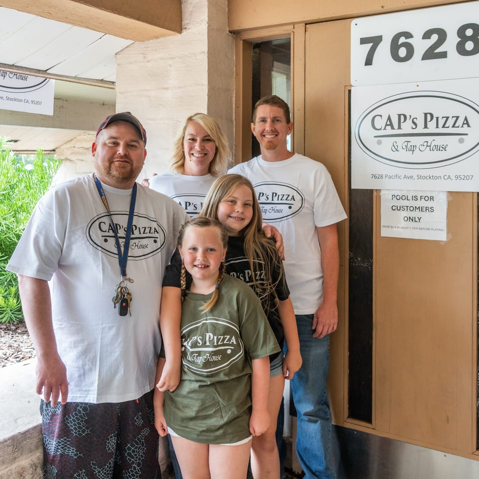 Two adults and two children wearing Cap's Pizza & Tap House shirts stand outside the restaurant.